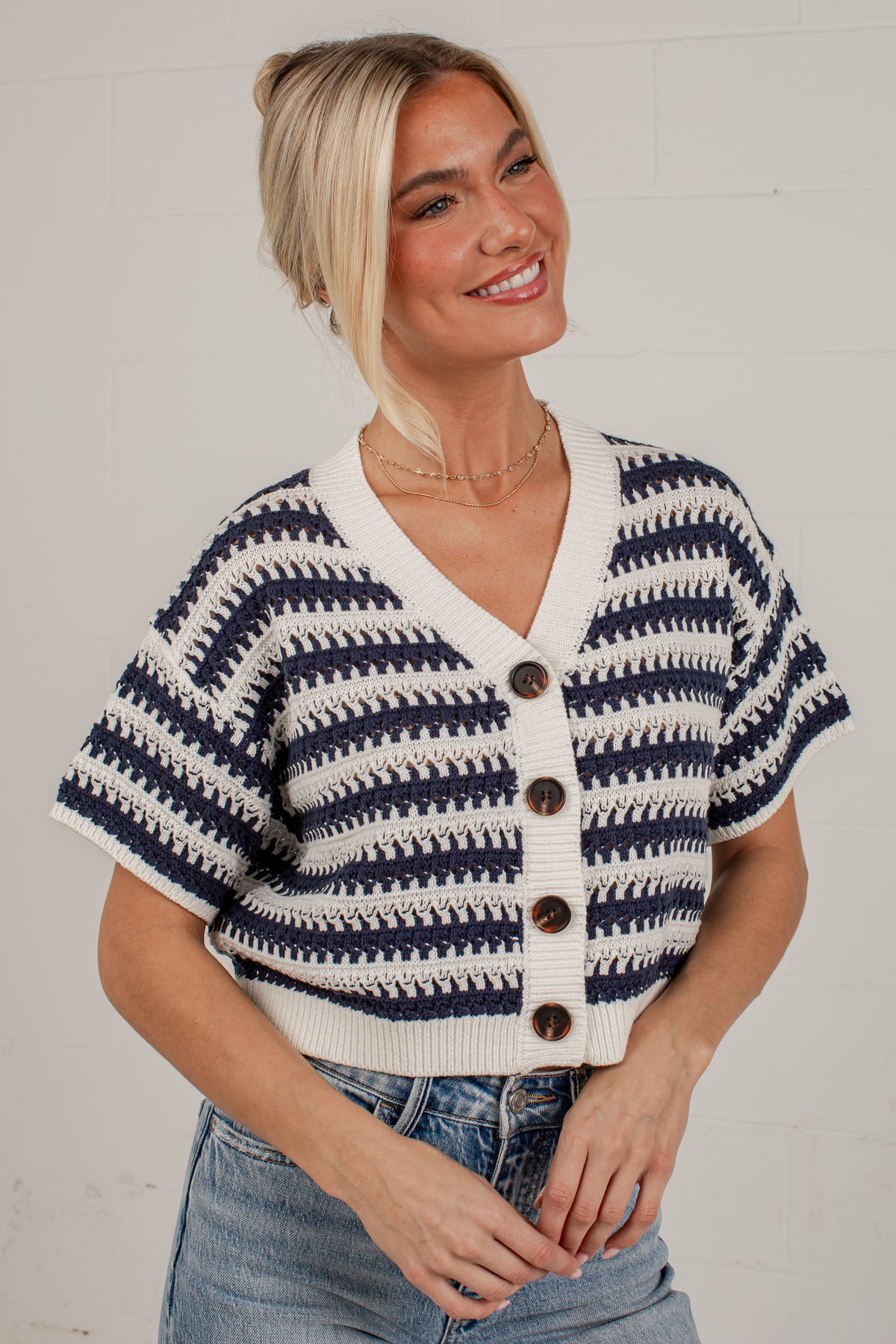 A woman wearing a short-sleeve knit cardigan with a navy and off-white horizontal striped pattern, brown buttons, and a V-neck, paired with blue jeans against a white brick wall.