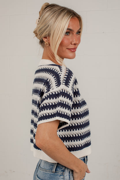 A woman wearing a navy blue and off-white patterned knit short-sleeve sweater, paired with blue jeans, standing against a light gray brick wall.