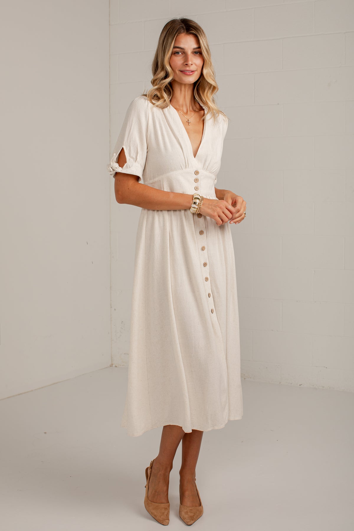 A woman modeling a cream-colored midi dress with a wrap-style V-neckline, button-down front, and cutout sleeves, standing against a plain white studio wall.