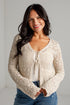 A smiling woman with long dark hair wears a cream-colored, long-sleeve crochet knit cardigan over a white top and blue jeans, standing against a white brick wall.