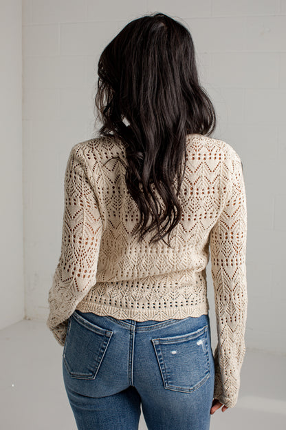 A woman viewed from behind wearing a cream-colored, long-sleeve open-knit sweater and blue jeans against a white brick wall.