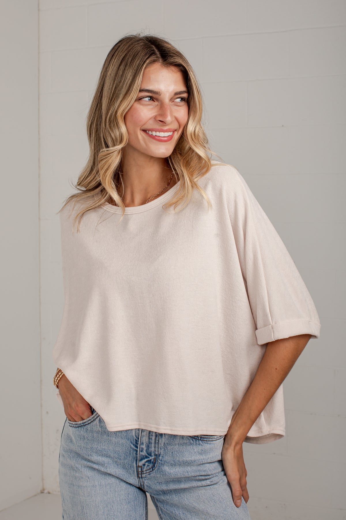 A woman wearing a light beige, oversized short-sleeve top with rolled cuffs and light wash blue jeans, standing against a light gray wall.