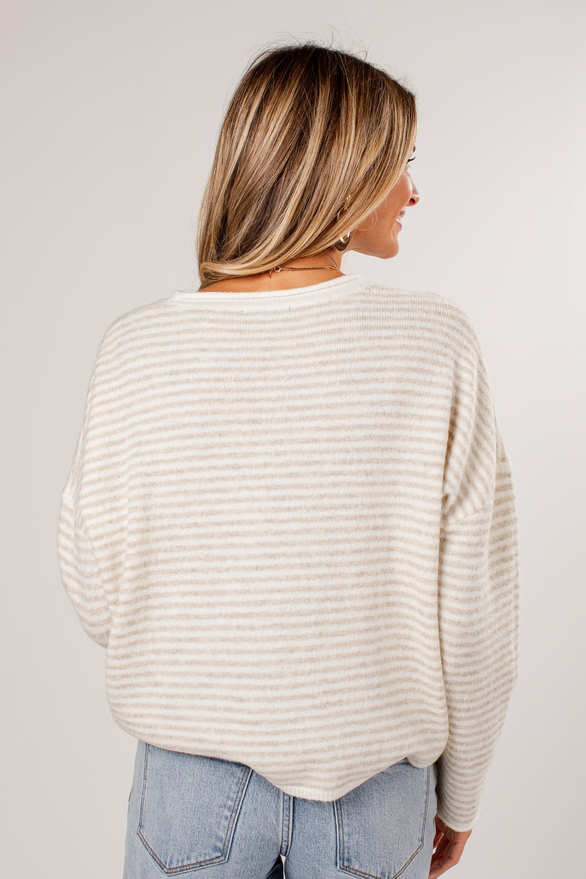 A woman shown from the back wearing a cream and beige horizontally striped knit sweater and light blue jeans, photographed against a plain light gray studio background.