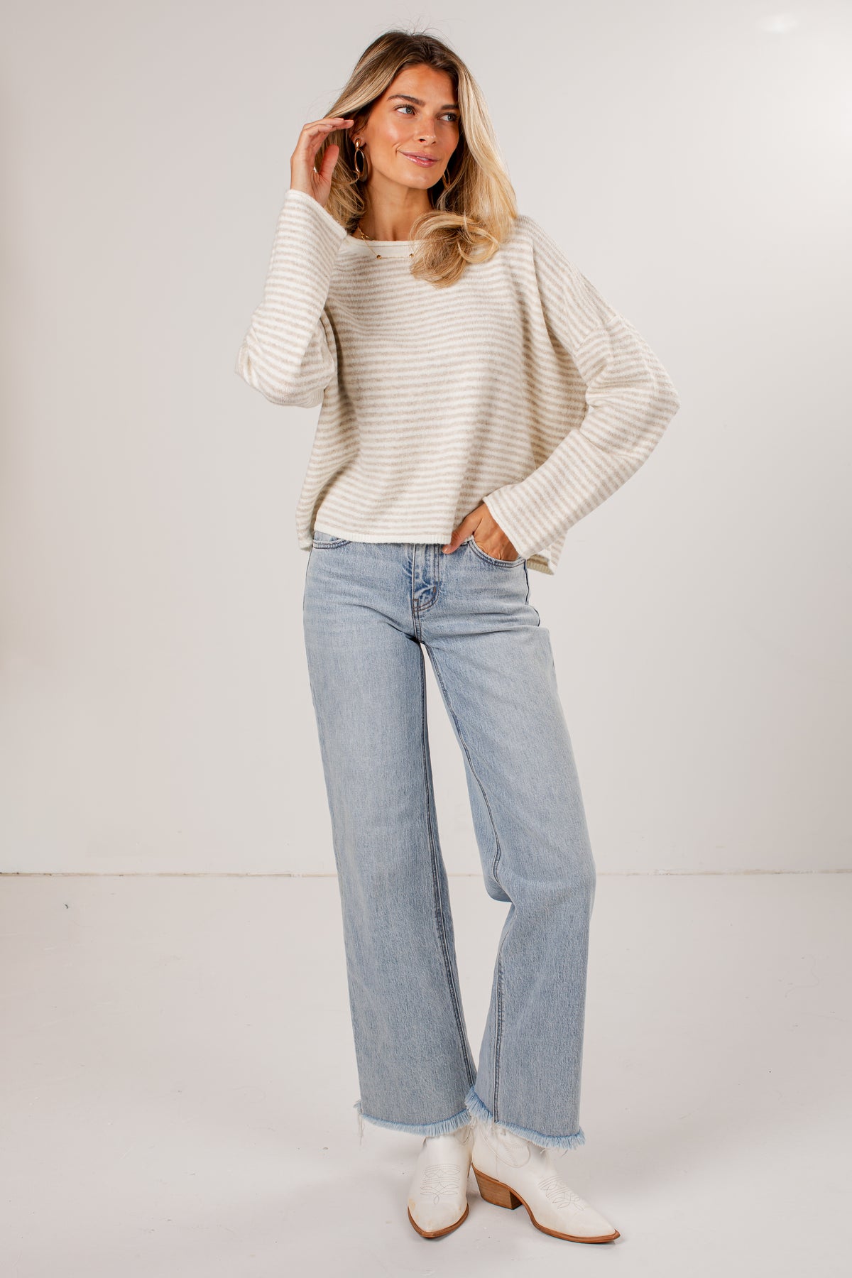 A woman modeling a cream and white horizontally striped knit sweater, light-wash wide-leg jeans, and white cowboy boots against a plain white studio background.