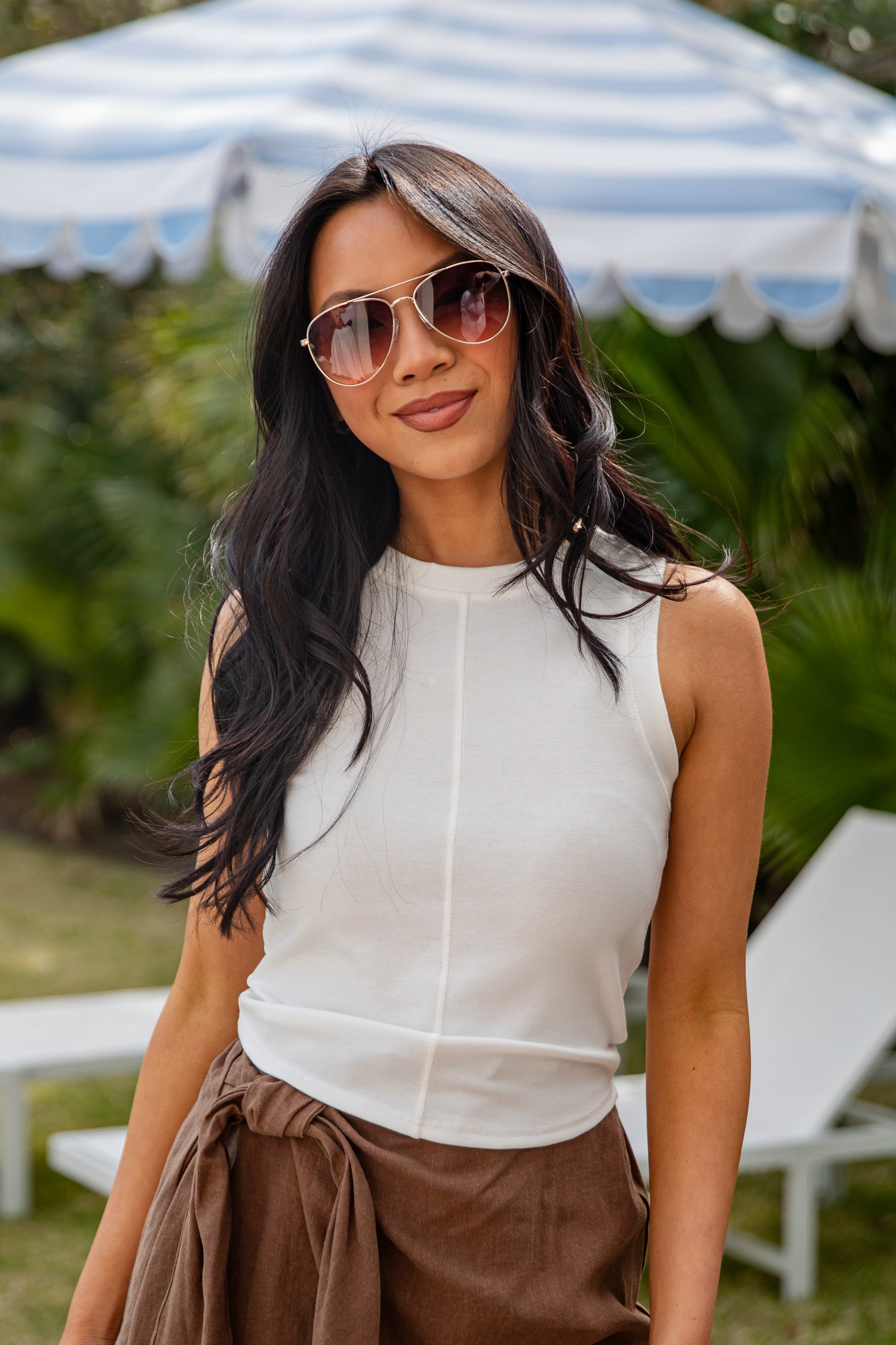 Woman wearing sunglasses and a white sleeveless top with brown pants outdoors.