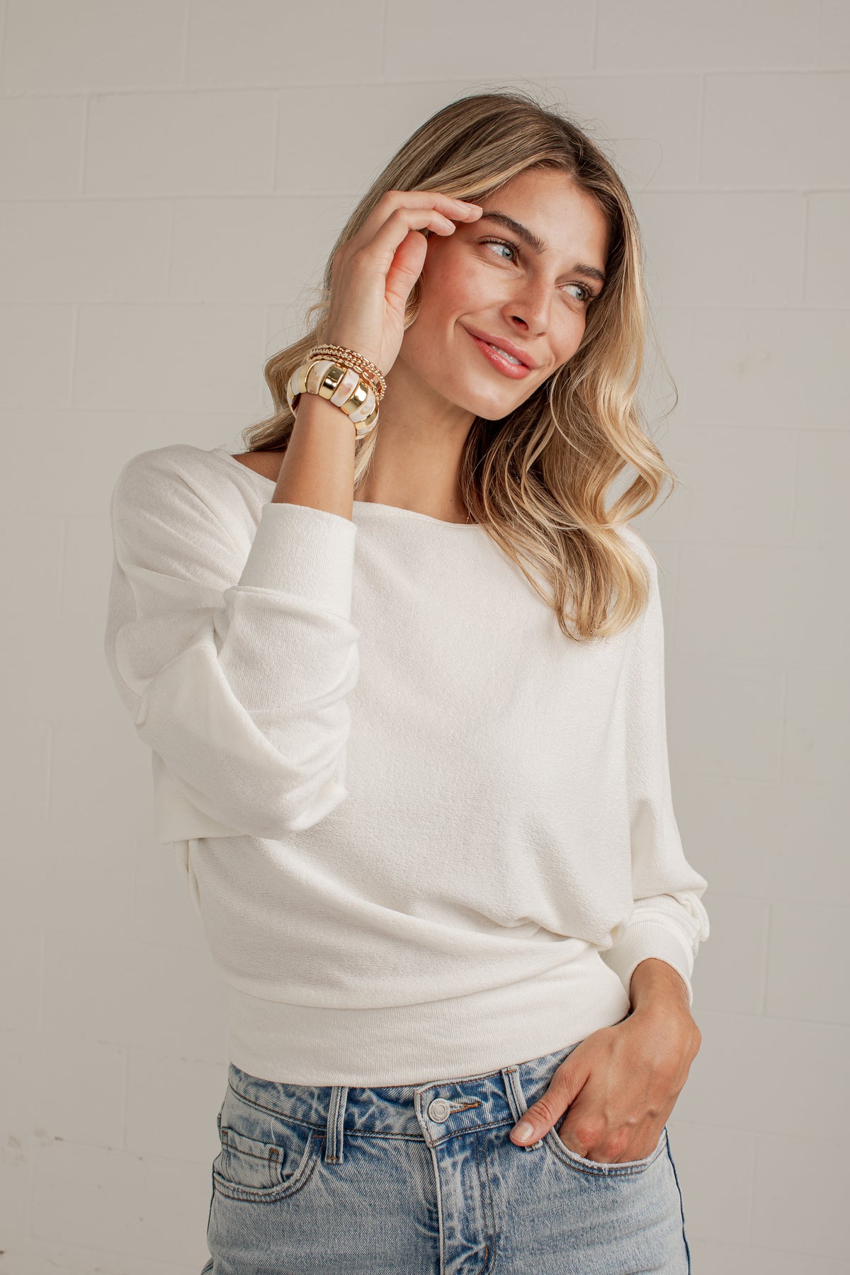 A woman wearing a white long-sleeve knit sweater and light-wash blue jeans, standing against a light gray brick wall.
