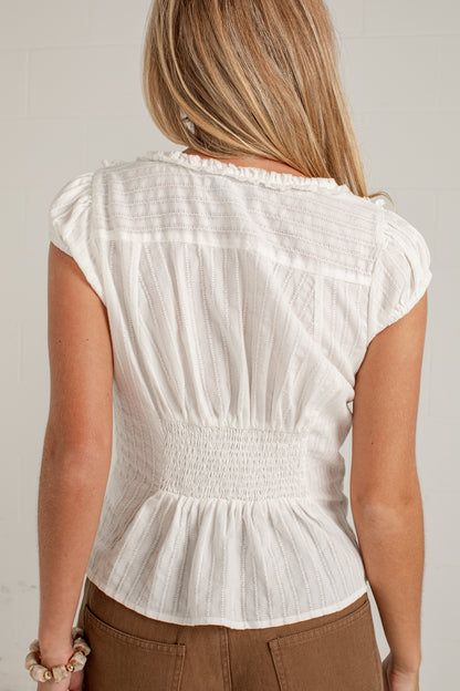 A woman viewed from the back wearing a white textured short-sleeve blouse with a smocked waist and ruffled neckline, paired with brown pants.
