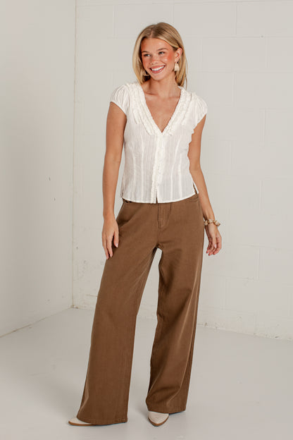 A woman modeling a white short-sleeve button-front blouse with pleated detailing and wide-leg brown pants, standing against a white studio wall.