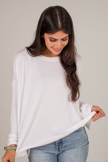 A woman wearing a white long-sleeve crewneck top and light blue jeans, holding up the hem of the shirt against a plain light gray background.