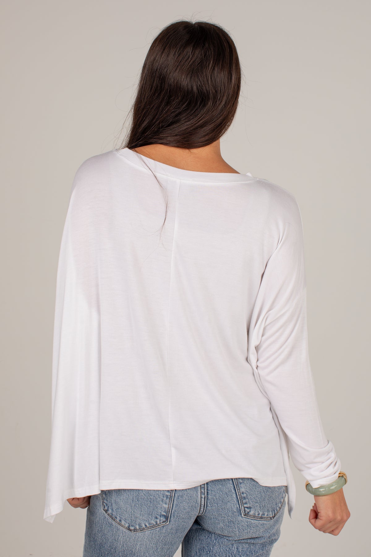 A woman viewed from the back wearing a white long-sleeve top and light blue jeans against a plain light gray studio background.