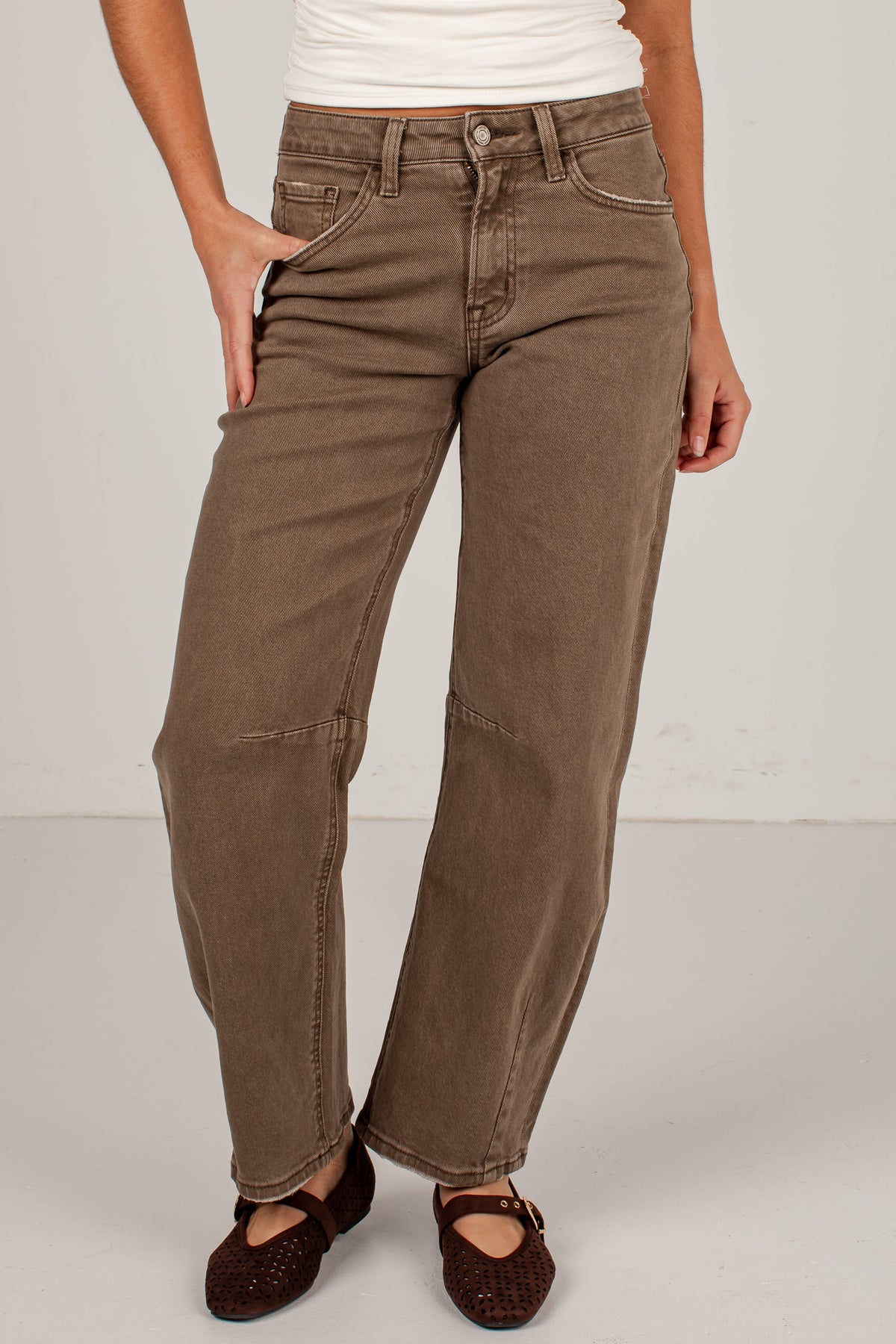 A person wearing brown wide-leg jeans with a white ribbed tank top and dark brown flat shoes, standing against a light gray studio background.