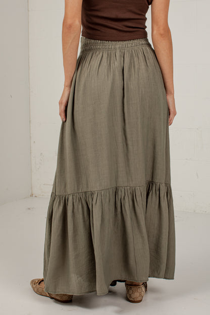 A woman wearing a long, tiered maxi skirt in a muted sage green color, paired with a brown top and cork sandals, standing against a white studio wall.