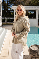 Woman in a beige shirt and white pants standing by a pool with trees in the background