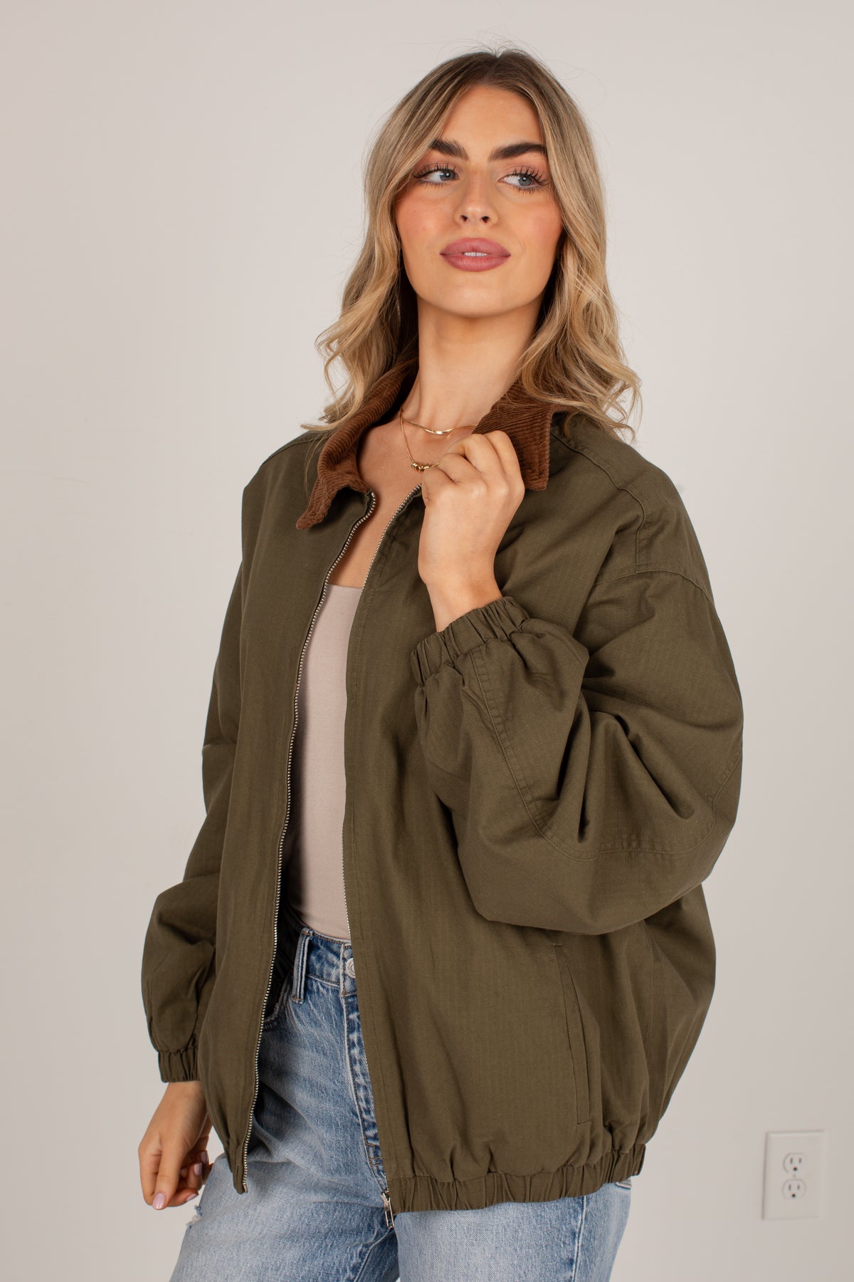 A woman wearing an oversized olive green utility jacket with a brown collar, styled over a beige top and blue jeans, standing against a plain light gray wall.