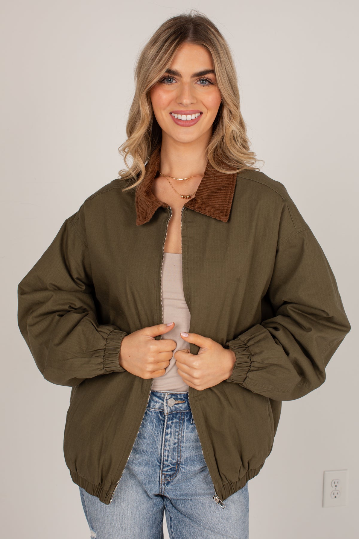 A woman wearing an olive green bomber jacket with a brown corduroy collar, styled over a beige top and blue jeans, standing against a plain light gray wall.