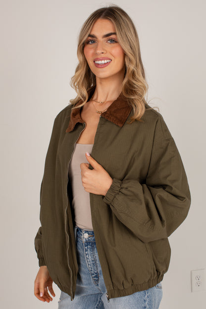 A woman wearing an olive green lightweight jacket with a brown corduroy collar, styled over a beige top and blue jeans, standing against a plain light gray wall.