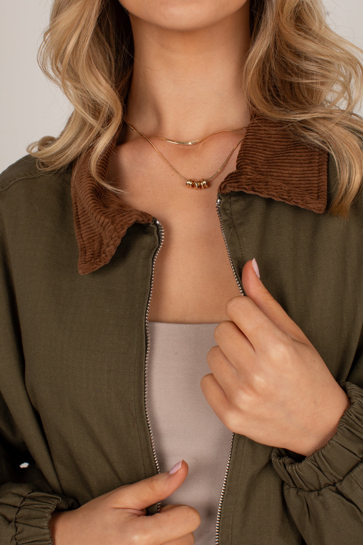 Close-up of a woman wearing an olive green zip-front jacket with a brown corduroy collar, layered over a light beige top. She is also wearing delicate gold necklaces.