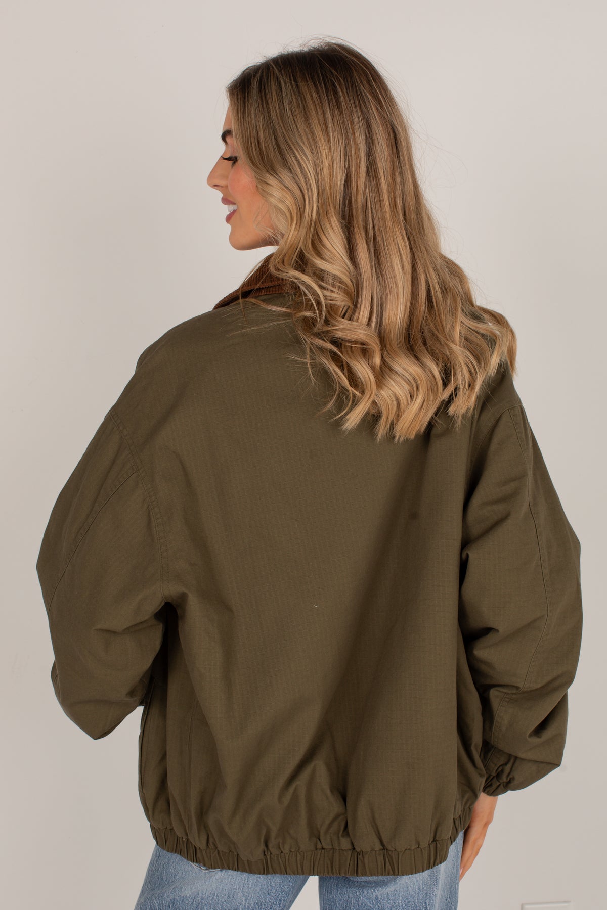 A woman with blonde wavy hair is shown from the back wearing an olive green bomber jacket and light blue jeans, photographed against a plain light gray studio background.