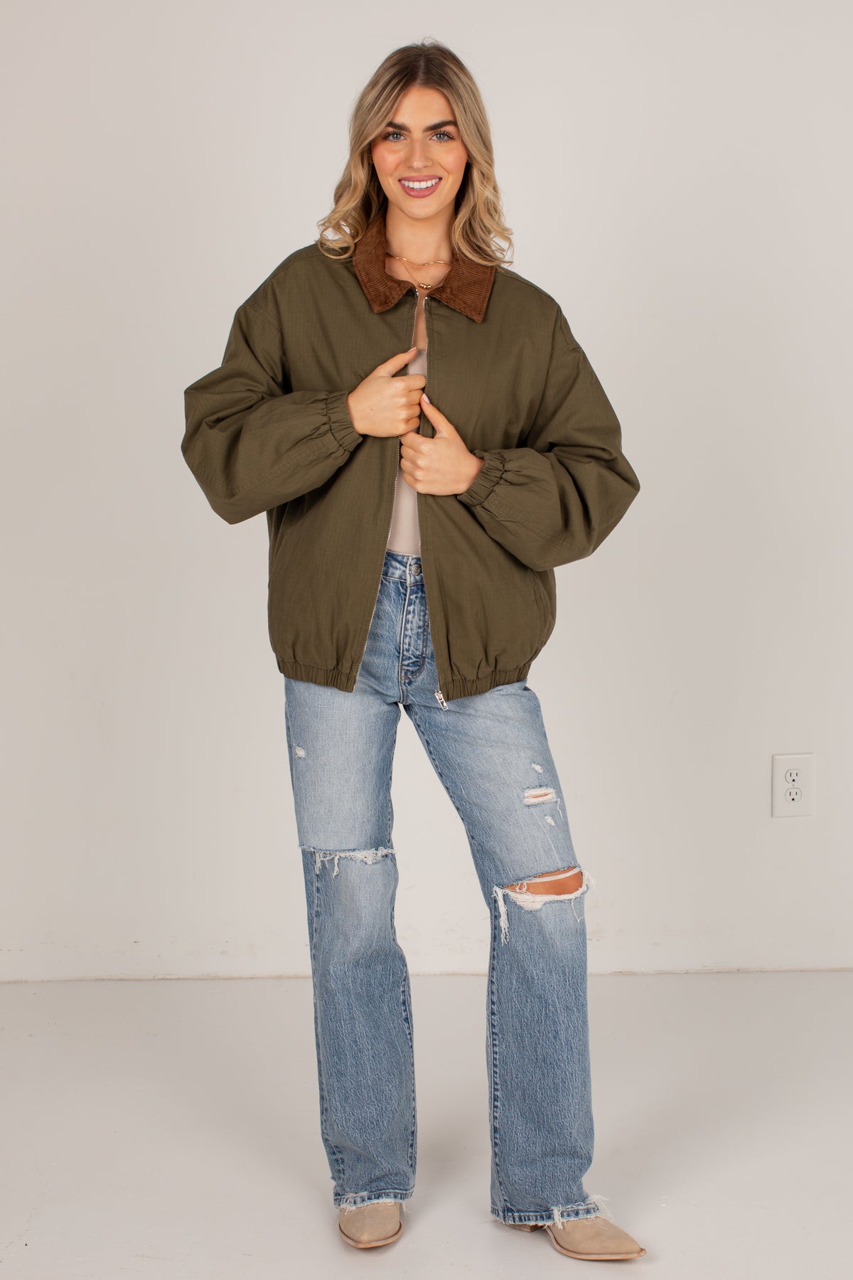 A woman modeling an olive green bomber jacket with a brown collar, paired with distressed light blue jeans and beige shoes, standing against a plain light gray studio background.