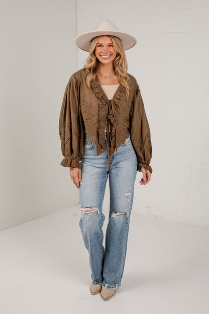 A woman modeling a brown ruffled long-sleeve blouse with a tie waist, paired with light-wash distressed jeans, beige heels, and a wide-brim beige hat, standing against a white studio wall.