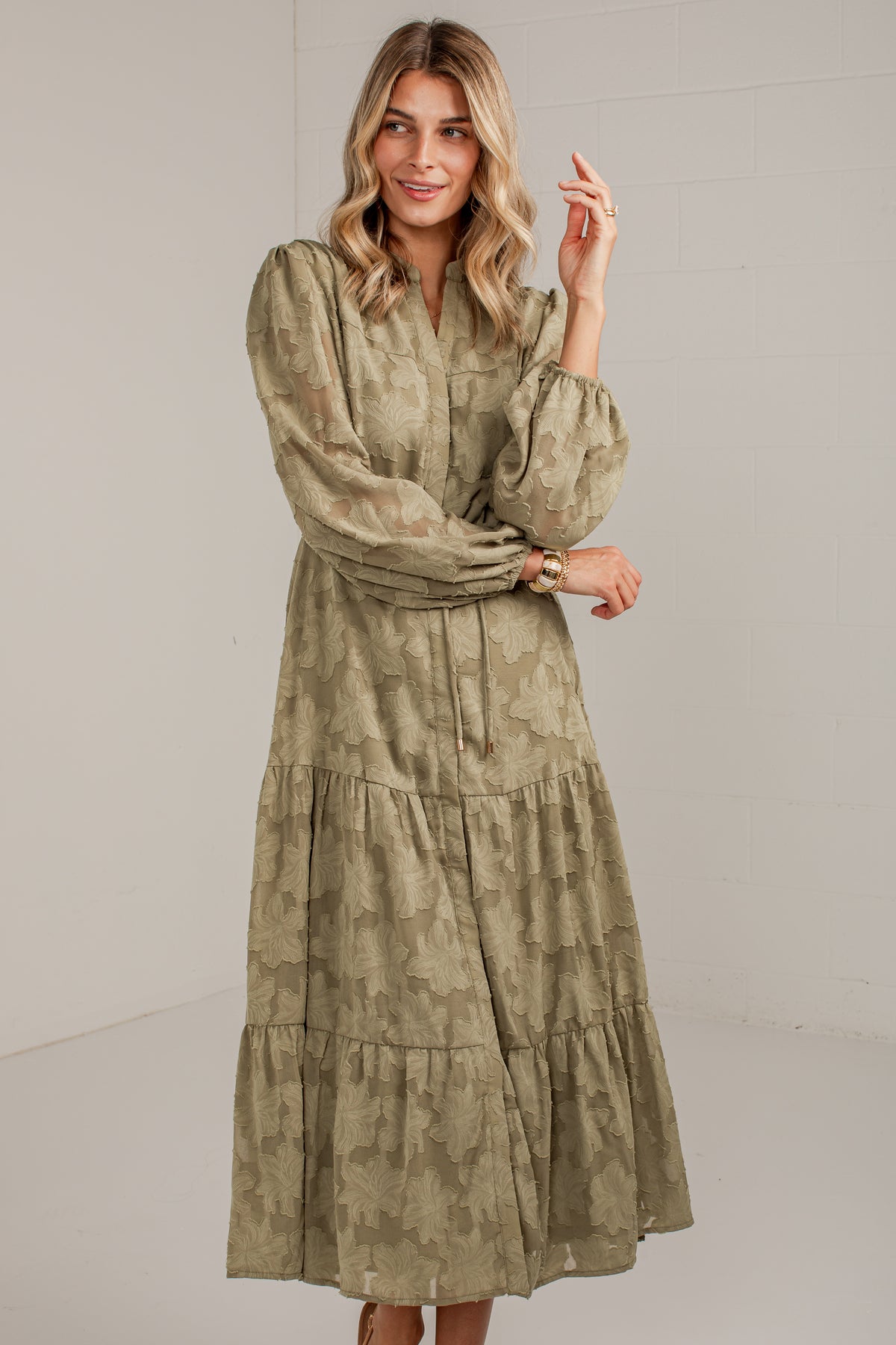 A woman modeling a long-sleeved, tiered midi dress with a muted olive green and beige abstract pattern, set against a plain light gray studio background.