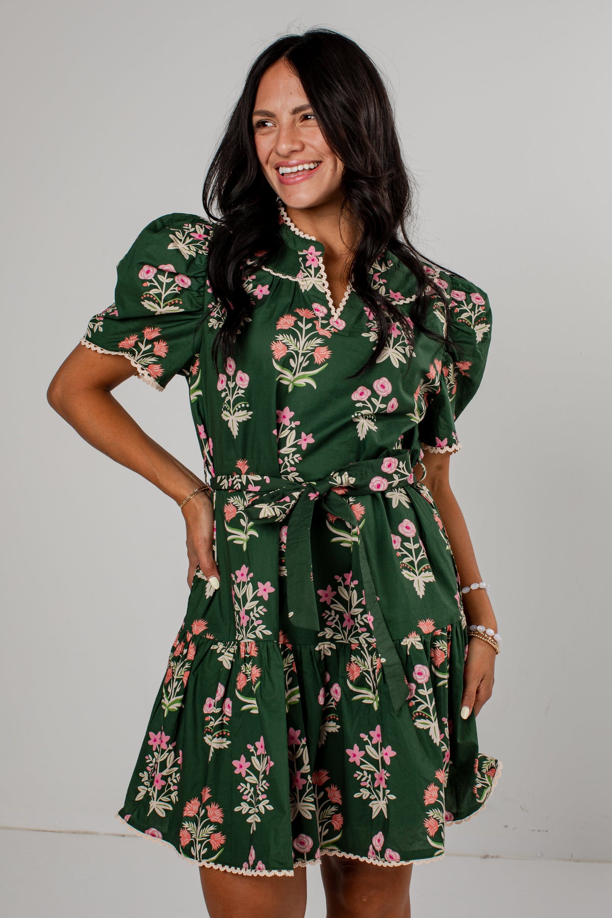 A woman modeling a dark green short-sleeve mini dress with a pink and white floral print, featuring puff sleeves, a collar, and a matching fabric tie belt, set against a plain light gray background.