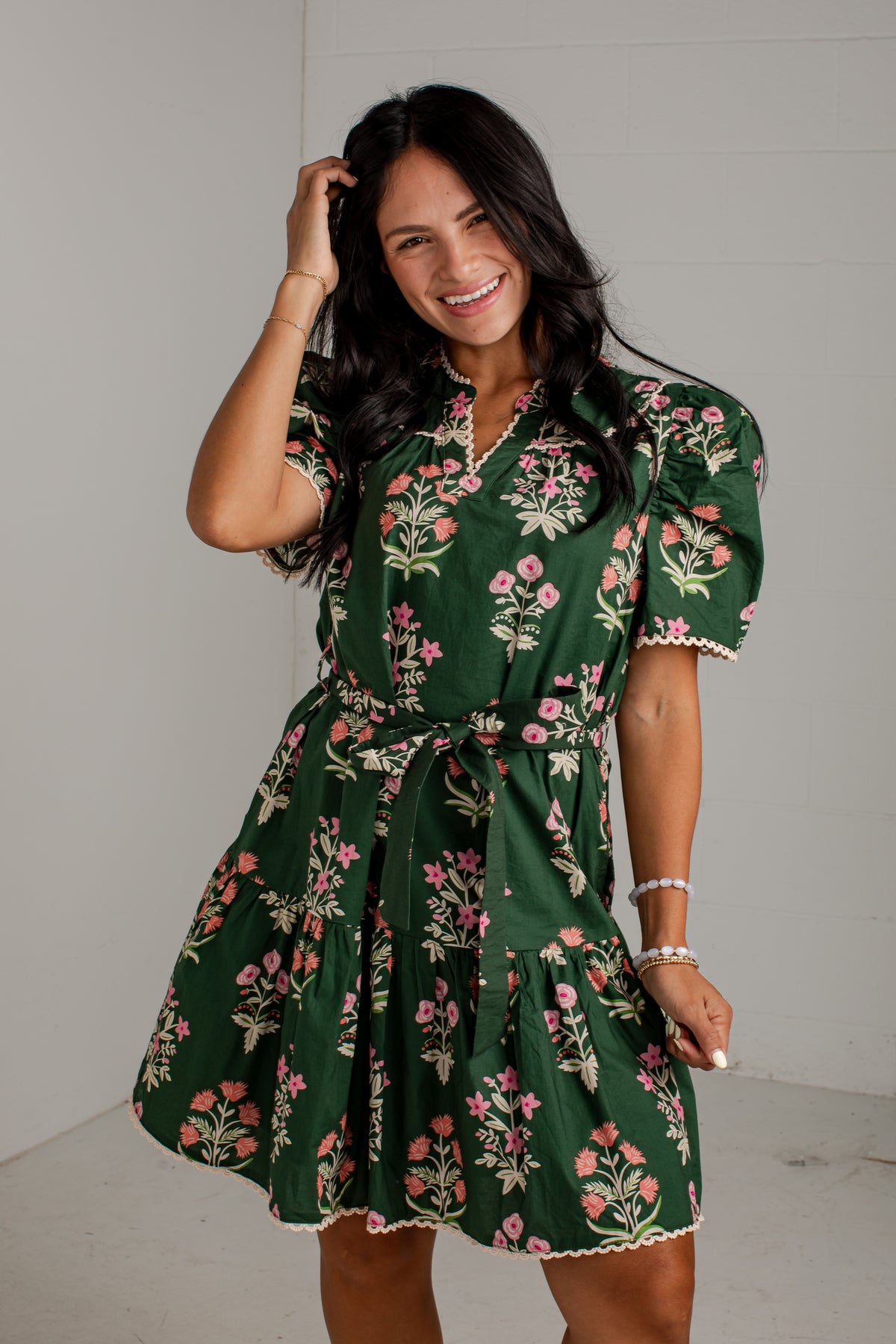 A woman modeling a dark green short-sleeve floral dress with pink and white flowers, a tie waist, and ruffled trim, standing against a light gray wall.