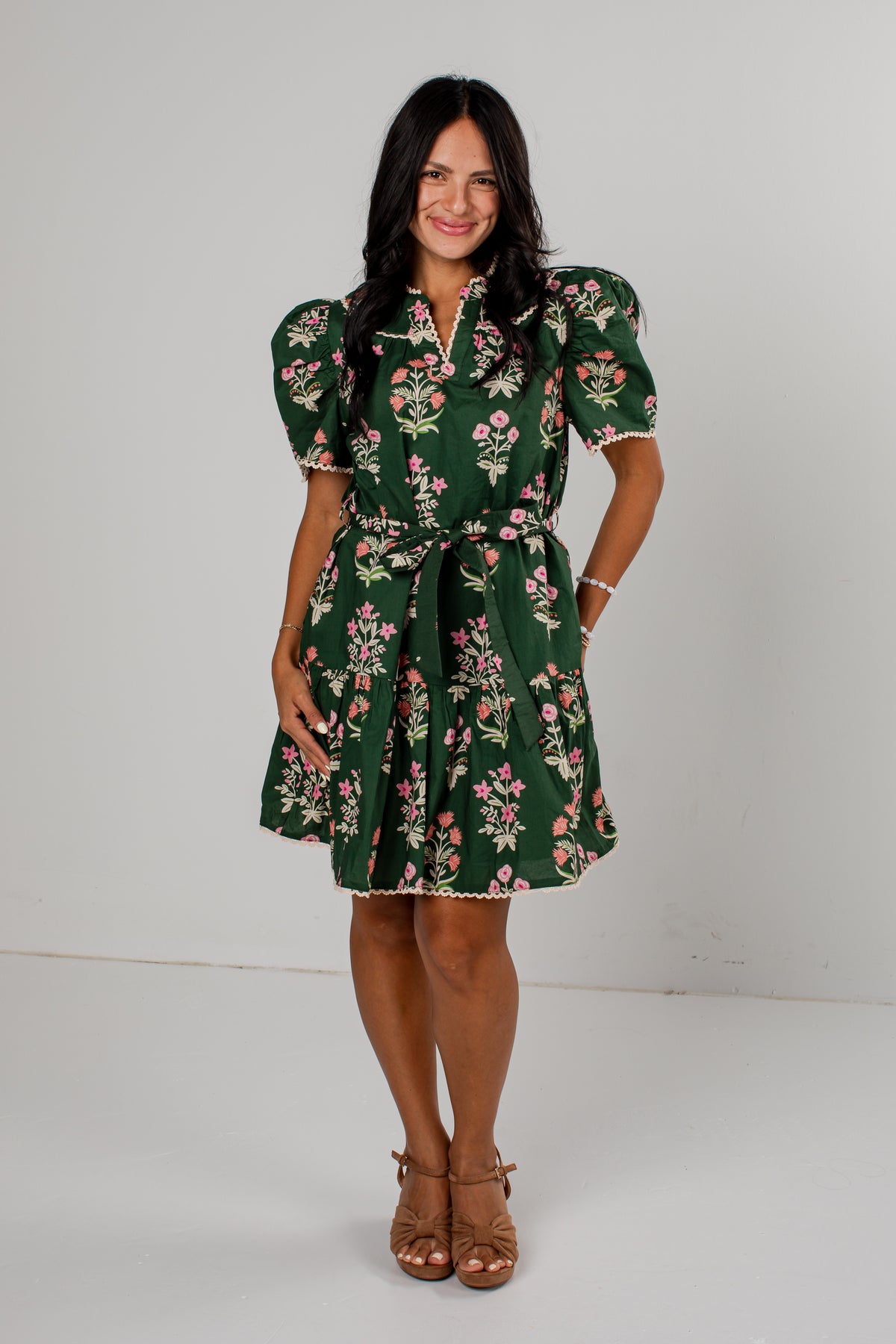 A woman modeling a dark green short-sleeve mini dress with a pink and white floral print, puff sleeves, and a matching tie belt, standing against a plain light gray studio background.