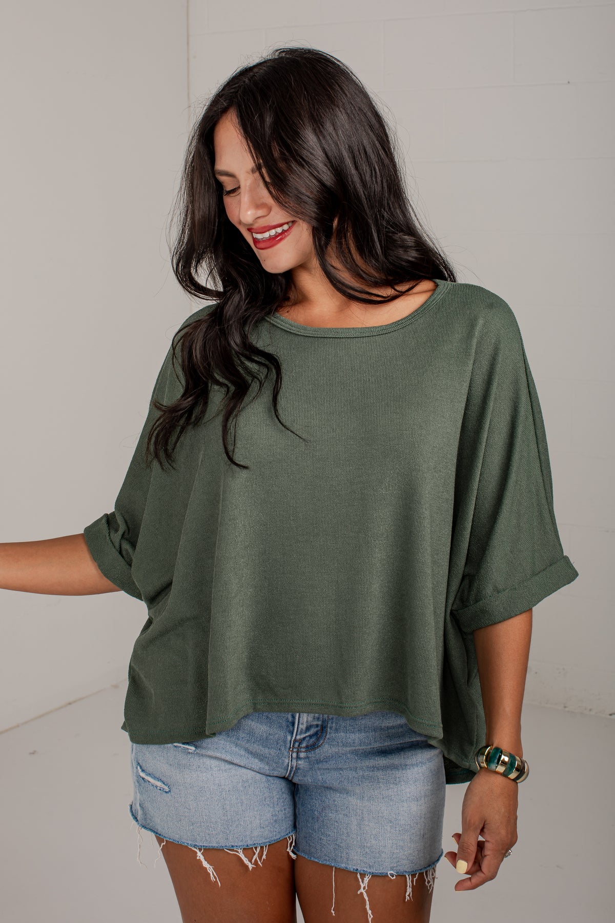 A woman wearing a loose, short-sleeve olive green top and light-wash denim shorts, standing against a plain light gray wall.