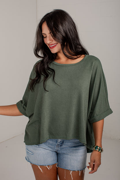 A woman wearing a loose, short-sleeve olive green top and light-wash denim shorts, standing against a plain light gray wall.
