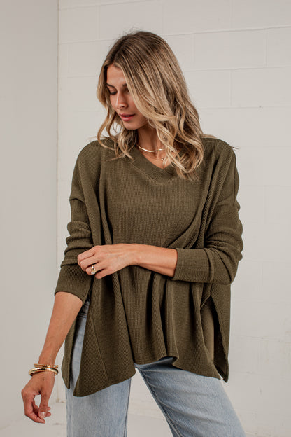 A woman wearing an olive green long-sleeve knit top with a draped, oversized fit and light-wash blue jeans, standing against a white brick wall.
