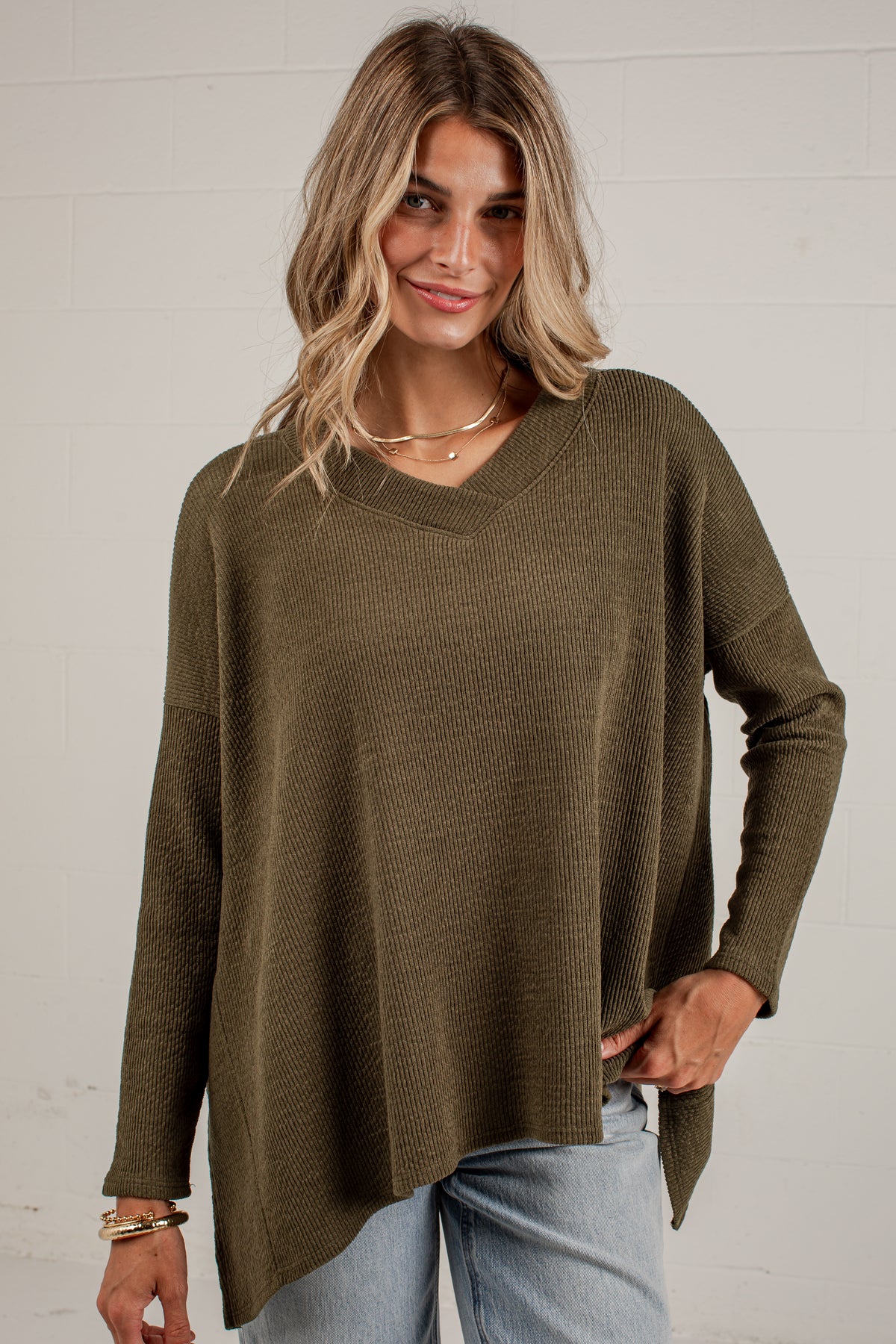 A woman wearing an olive green ribbed knit long-sleeve sweater with a V-neck and an asymmetrical hem, paired with light blue jeans, standing against a white brick wall.