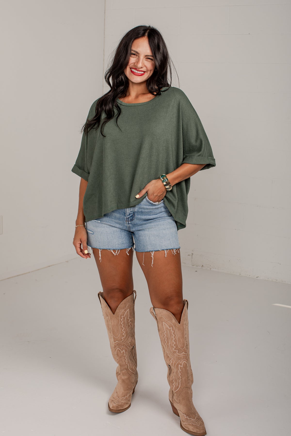 A woman modeling a loose-fitting sage green short-sleeve top, paired with light-wash denim shorts and tan cowboy boots, standing against a plain white studio background.
