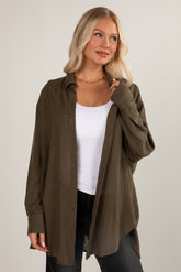 Woman wearing an olive green shirt over a white top and black pants on a plain background