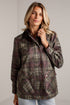 A woman wearing a brown and green plaid waxed jacket with dark brown corduroy collar and front buttons, paired with light blue jeans, standing against a white brick wall.