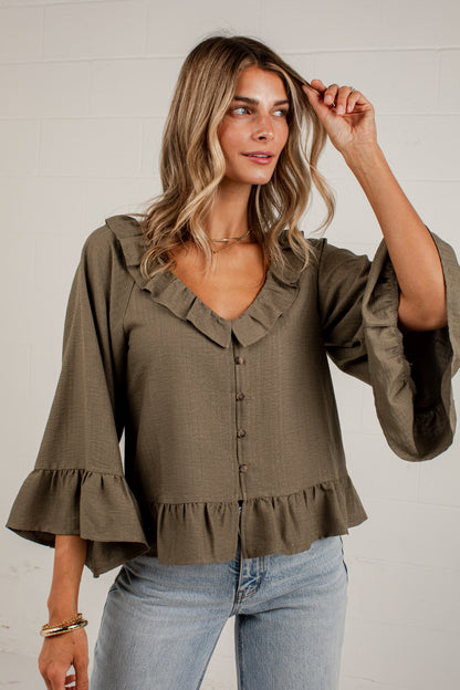 A woman wearing an olive green blouse with ruffled trim, button-front detail, and flared bell sleeves, paired with light blue jeans, standing against a white brick wall.