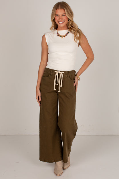 A woman modeling a white sleeveless top and olive green wide-leg drawstring pants against a plain light gray studio background.