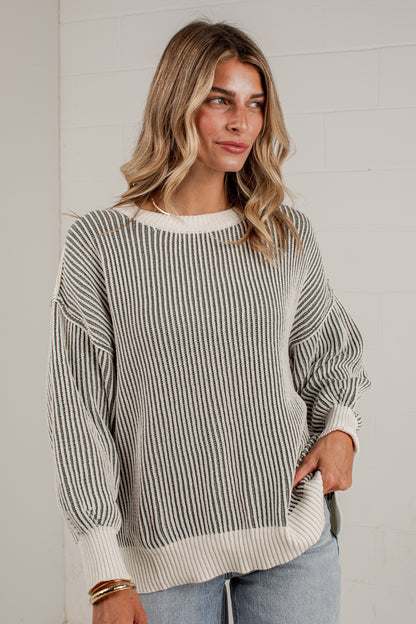 A woman wearing a long-sleeve knit sweater with vertical gray and off-white stripes, paired with light blue jeans, standing against a white paneled wall.