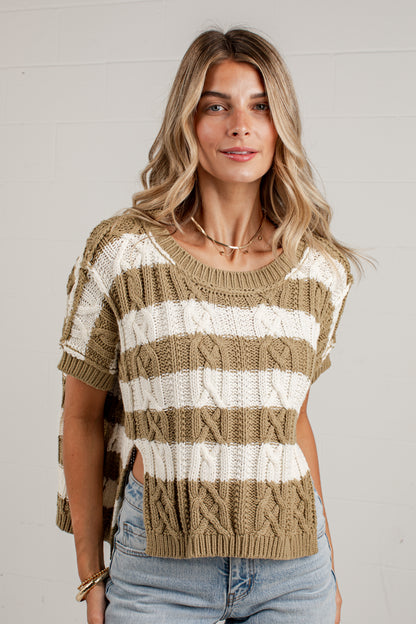 A woman wearing a short-sleeve knit sweater with horizontal beige and off-white stripes, paired with light-wash jeans, standing against a white paneled wall.