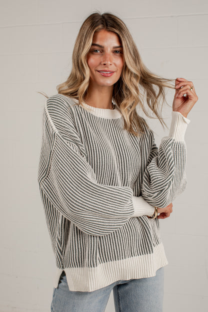 A woman wearing a cream and gray vertically striped knit sweater with balloon sleeves and ribbed cuffs, hem, and collar, paired with light blue jeans against a white paneled wall.