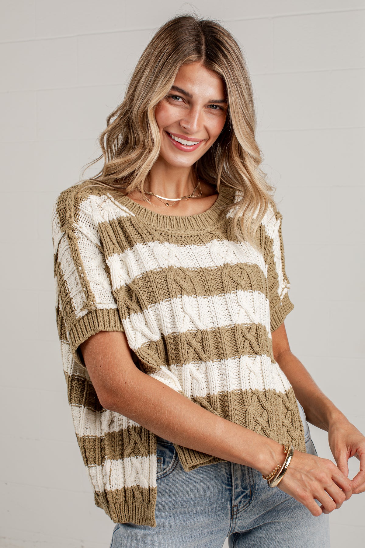 A woman wearing a short-sleeve knit sweater with horizontal stripes in tan and off-white, paired with light blue jeans, against a light gray paneled wall.