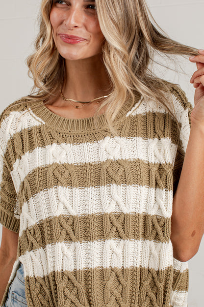 A woman wearing a short-sleeve knit sweater with horizontal stripes in taupe and off-white, featuring cable-knit patterns. The photo is a close-up crop of the upper body.