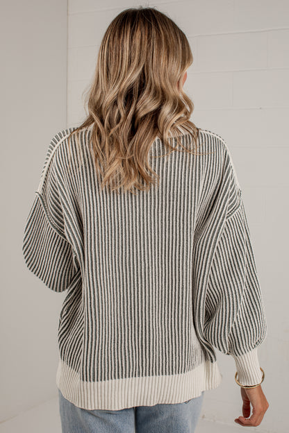 A woman viewed from behind wearing a cream and dark gray vertically striped knit sweater with balloon sleeves and ribbed trim, paired with light blue jeans against a white paneled wall.
