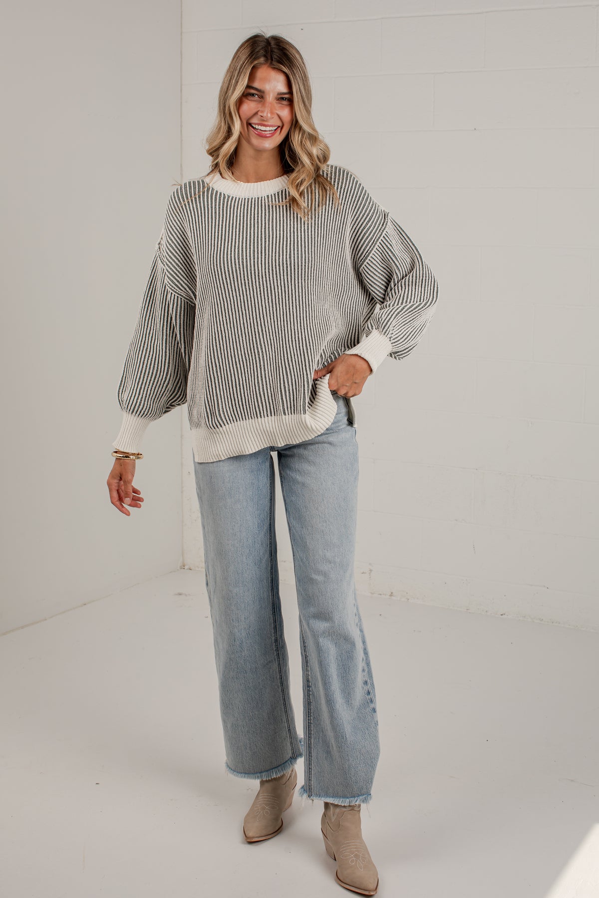 A woman modeling a ribbed knit sweater with wide sleeves, paired with light-wash wide-leg jeans and beige ankle boots, standing in a bright white studio.