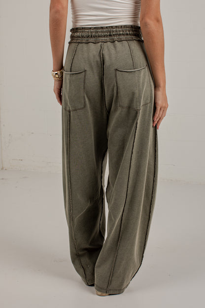 A person wearing loose, wide-leg olive green drawstring pants with large front patch pockets, paired with a white top and beige sneakers, standing against a plain light wall.
