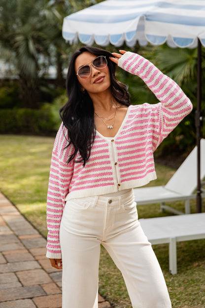 Woman wearing a pink and white striped cardigan and white pants outdoors.
