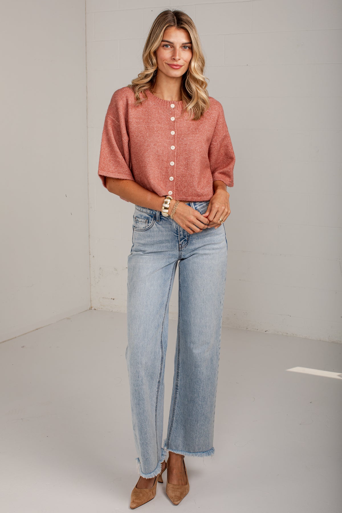A woman modeling a short-sleeve, button-front top in a dusty rose color paired with light-wash wide-leg jeans and tan heels, standing against a plain white studio wall.