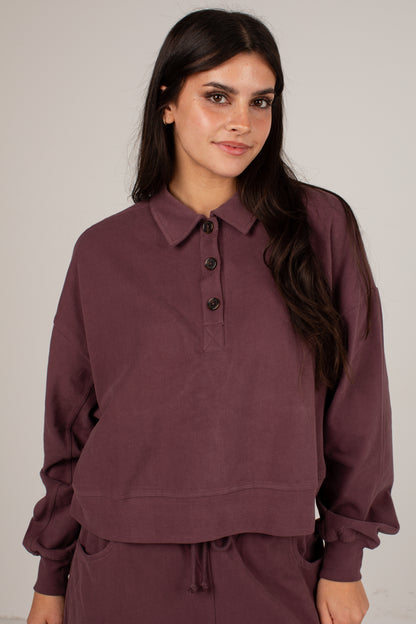 A woman modeling a dusty plum purple long-sleeve polo sweatshirt with a buttoned placket, paired with matching sweatpants against a neutral light gray background.