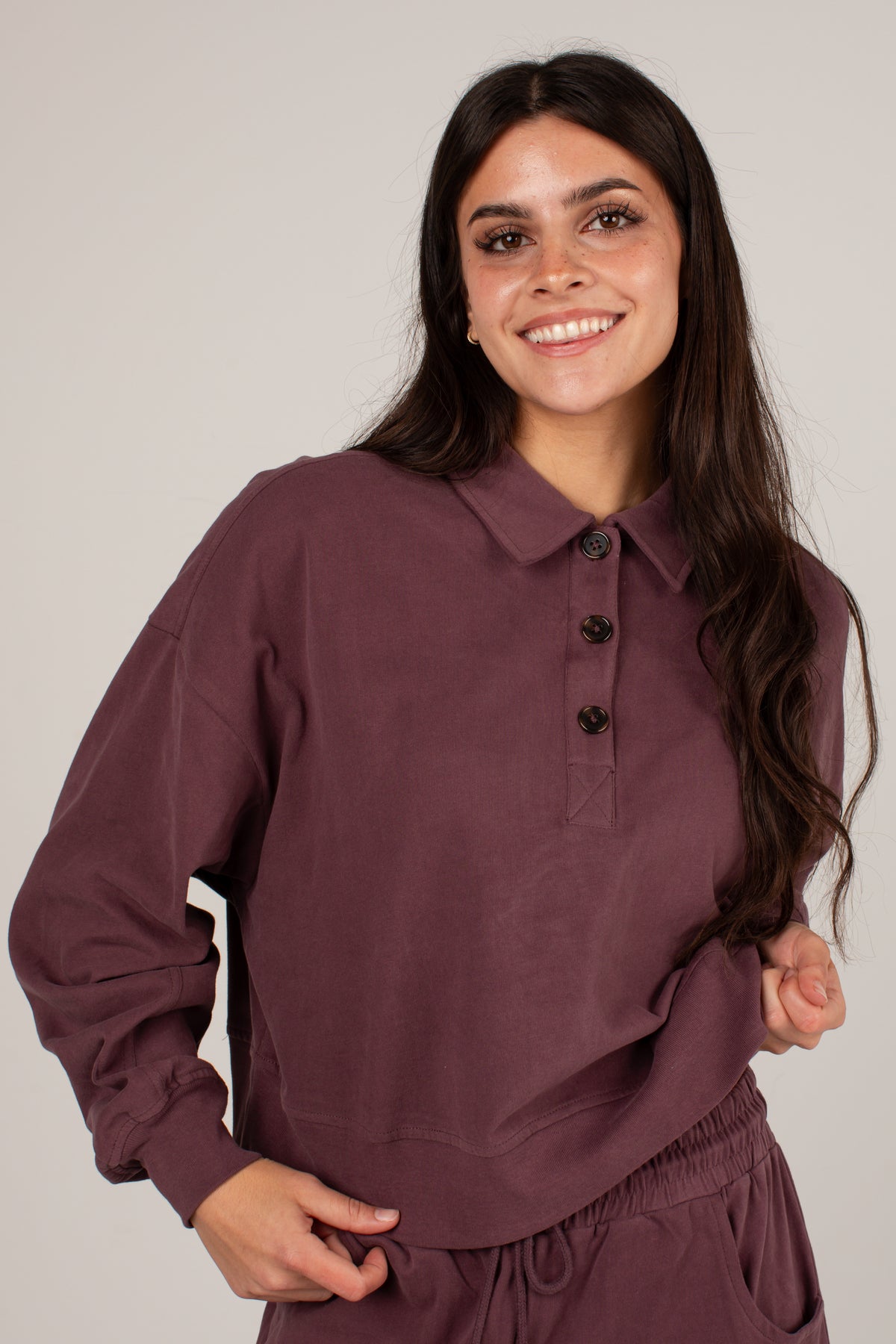 A woman wearing a long-sleeve polo shirt in a dusty plum purple color, paired with matching sweatpants, against a solid light gray background.