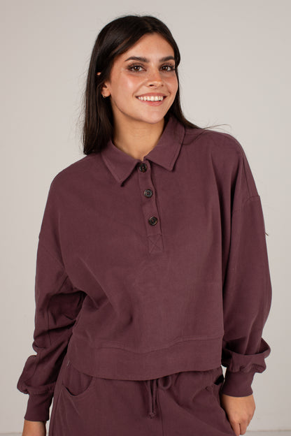 A woman wearing a dark plum-colored long-sleeve polo shirt with a buttoned collar, paired with matching pants, photographed against a neutral light gray background.